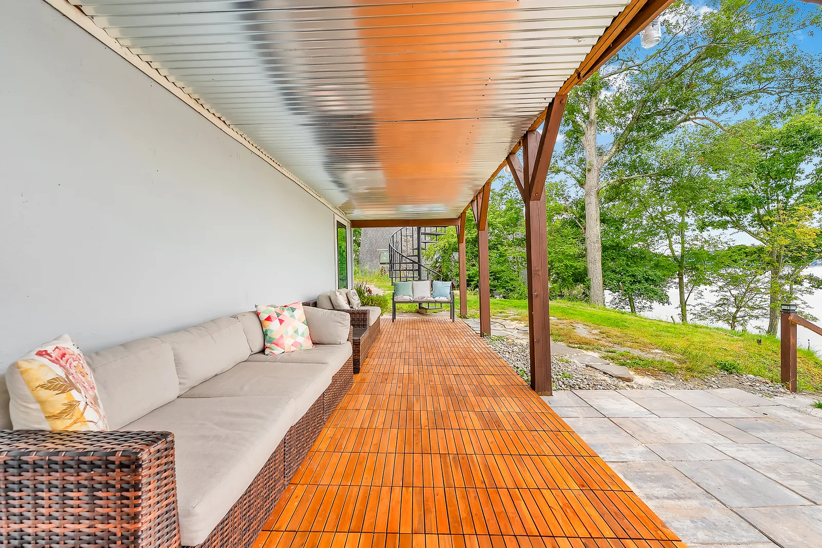 The covered porch down its length, a long cushioned bench on teak boards under a ribbed metal ceiling
