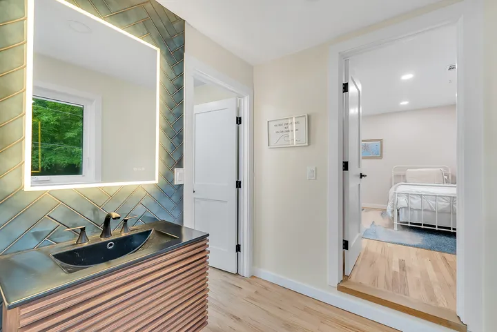 A bathroom vanity in dark herringbone tile under a lit mirror, an open door through to a bedroom beyond