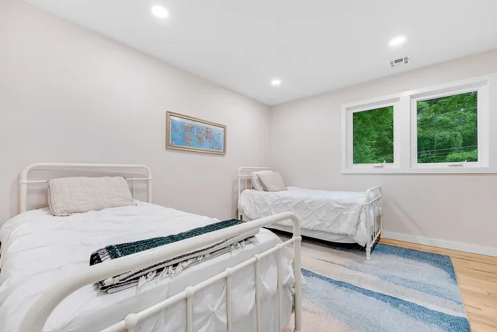 The Cozy Twin: two beds with white iron frames on a blue striped rug, a window onto the trees between them