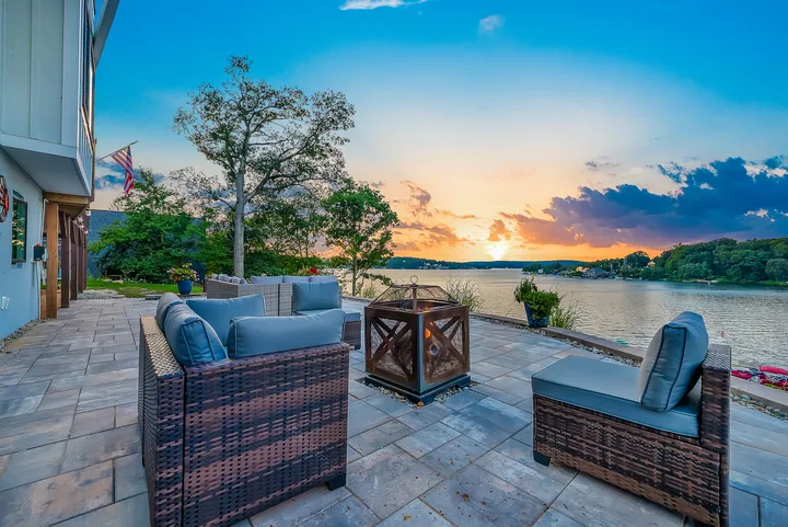 The stone patio at sunset, deep cushioned chairs around a low table, the sun going down over the far shore