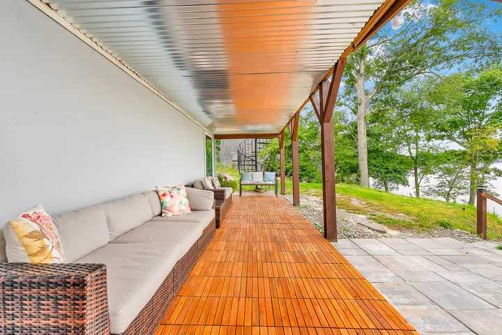 The covered porch down its length, a long cushioned bench on teak boards under a ribbed metal ceiling